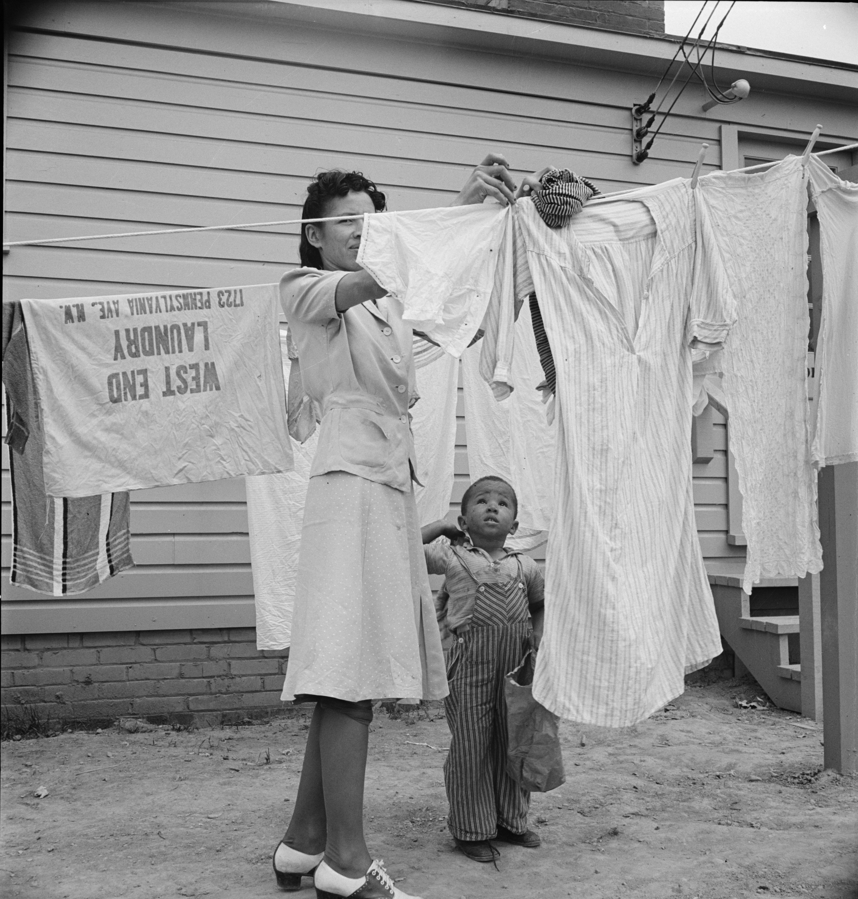 A woman hanging out washing with her young son in front of the community building in a FSA (Farm Security Administration) housing relief camp for African Americans. Arlington, Virginia, 1942. Source: Library of Congress.